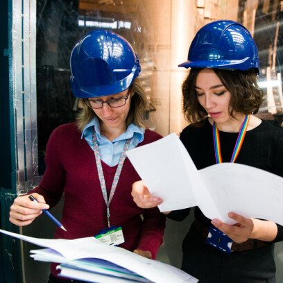 Two women wearing blue hardhats and lanyards reviewing documents