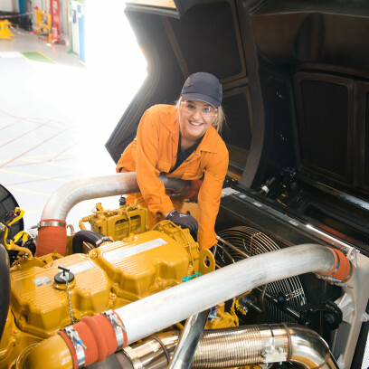A female wearing a black cap, safety goggles and orange overalls working on a large engine