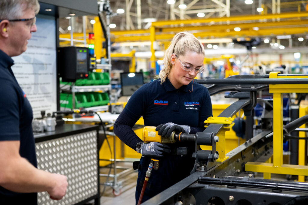 A student wearing safety goggles uses manufacturing equipment, observed by a teacher.