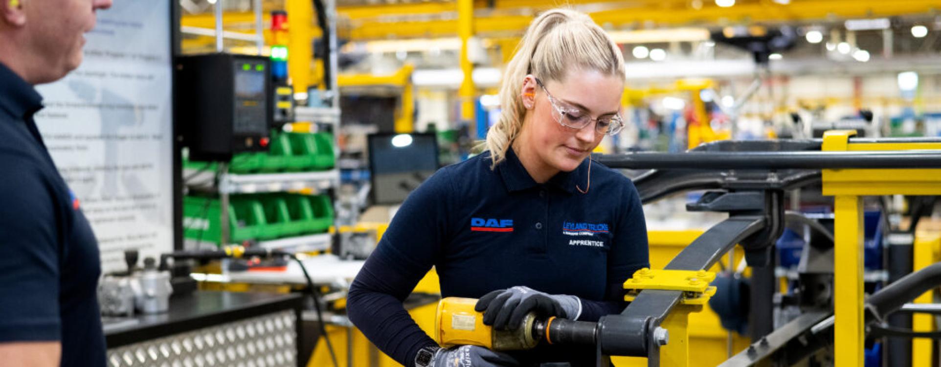 A student wearing safety goggles uses manufacturing equipment, observed by a teacher