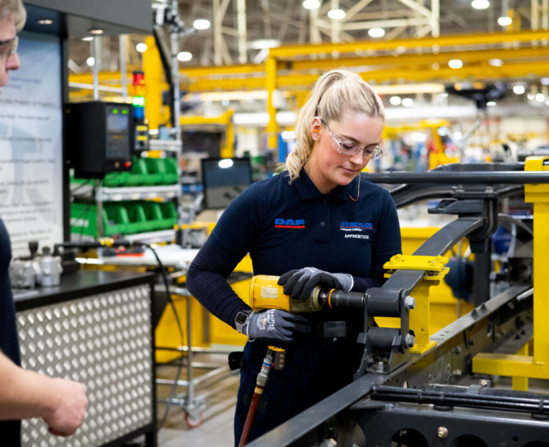 A student wearing safety goggles uses manufacturing equipment, observed by a teacher.