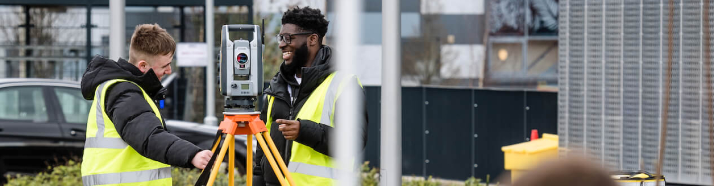 Two people wearing yellow safety vests are using surveying equipment outdoors.