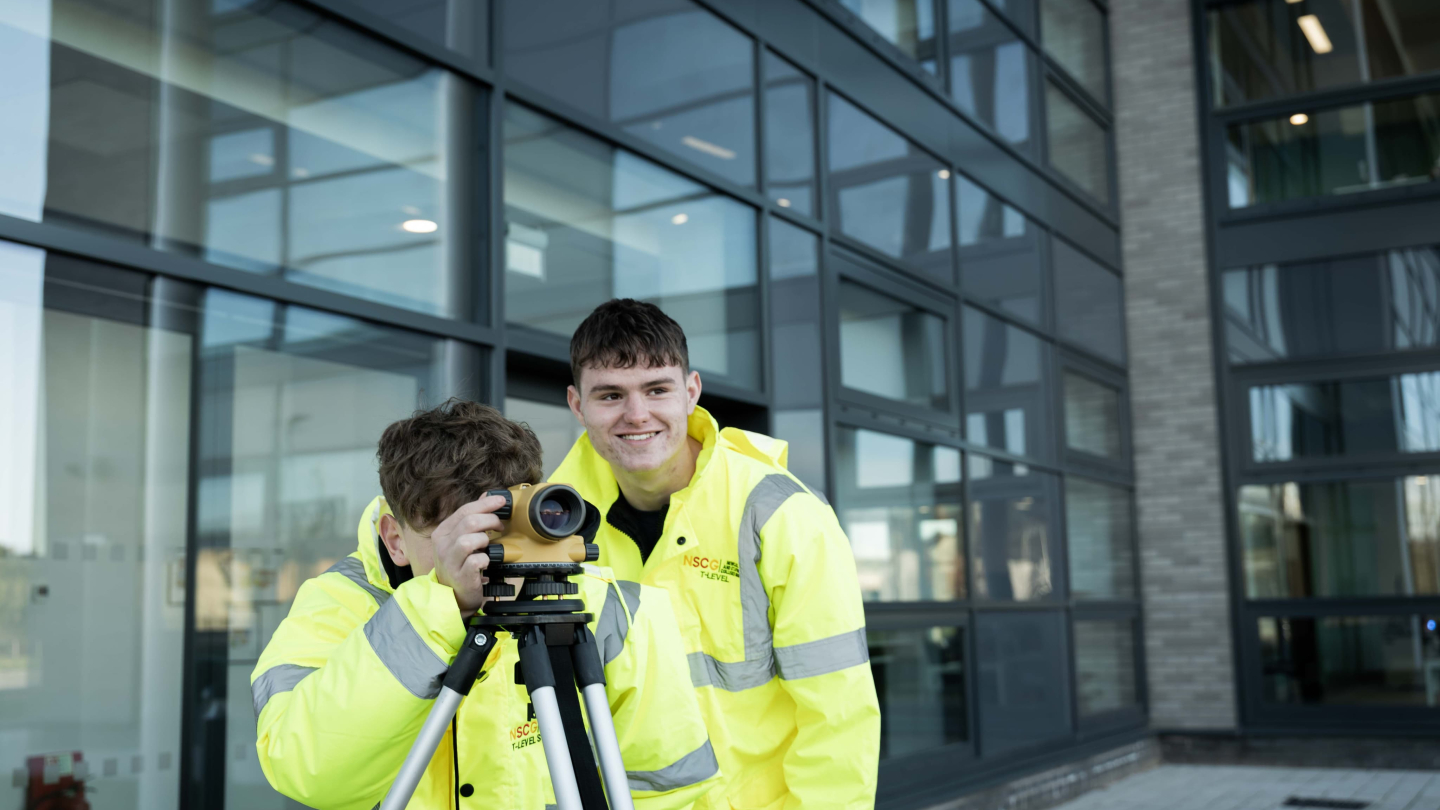 Two individuals in bright yellow jackets are working outdoors with a surveying instrument.