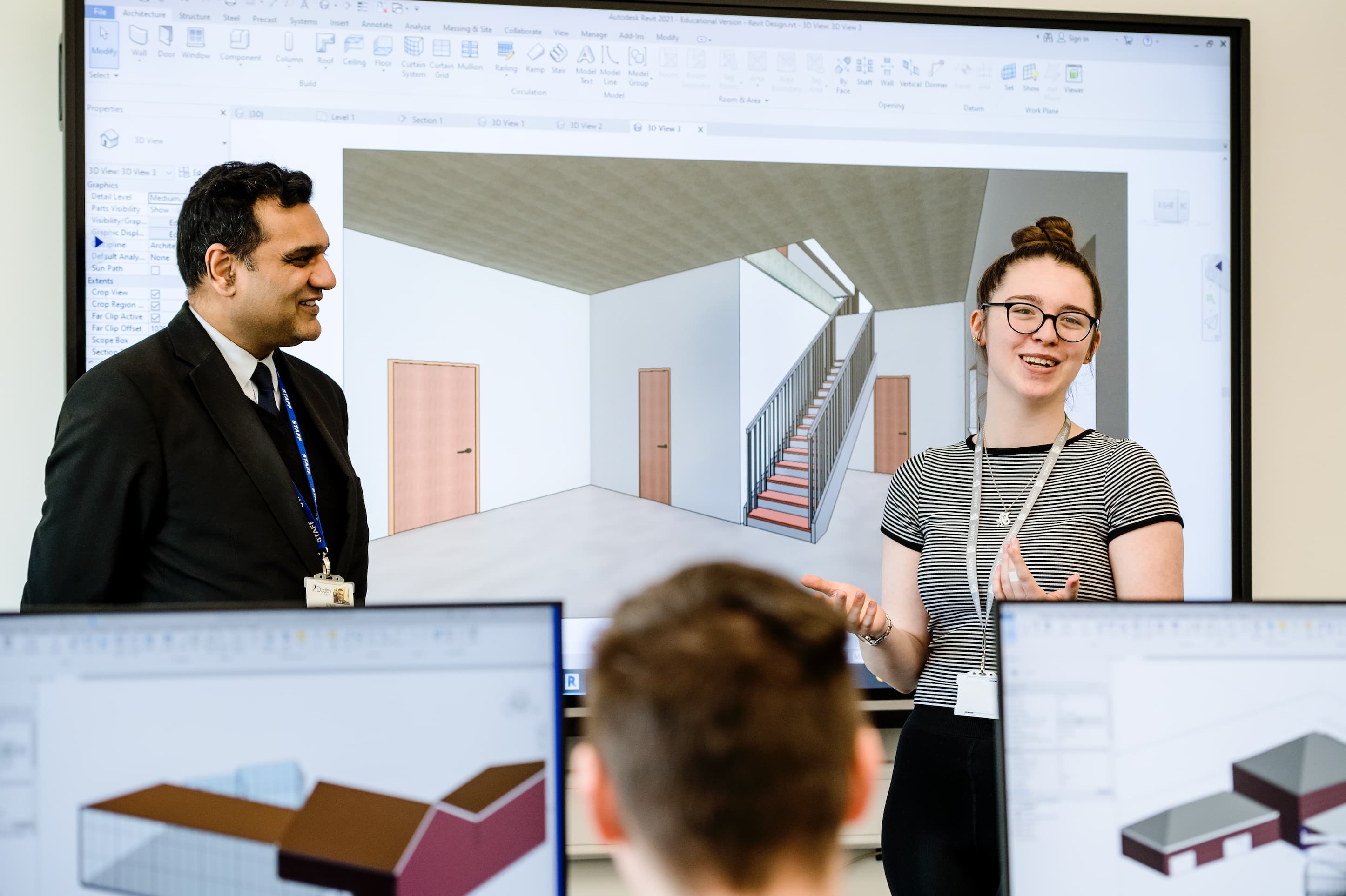 A person stands in front of a screen displaying architectural designs, gesturing as they speak to an audience.