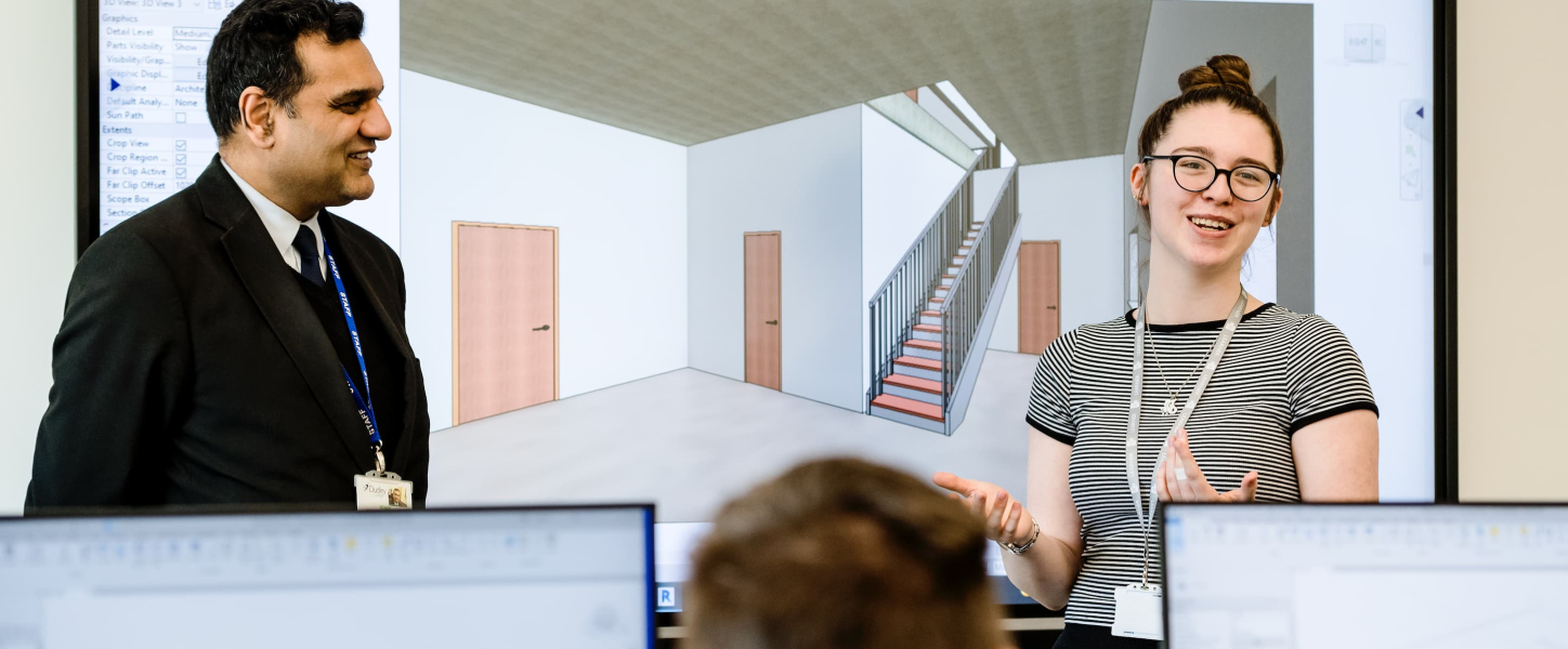 Two people presenting in front of a screen displaying a 3D model of an interior space.