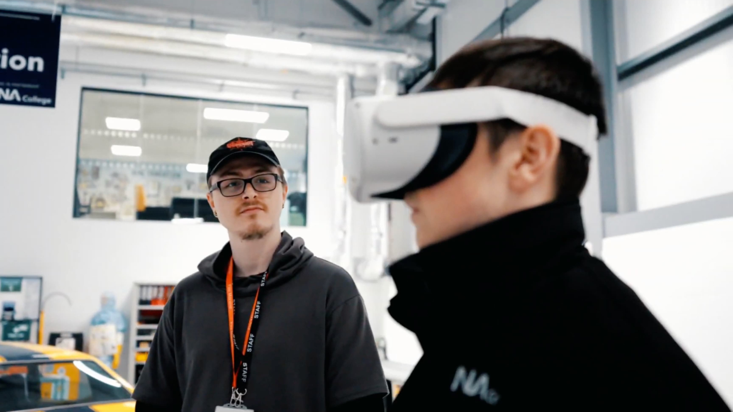 A student wearing a Virtual Reality headset stands next to an IoT staff member in a classroom.