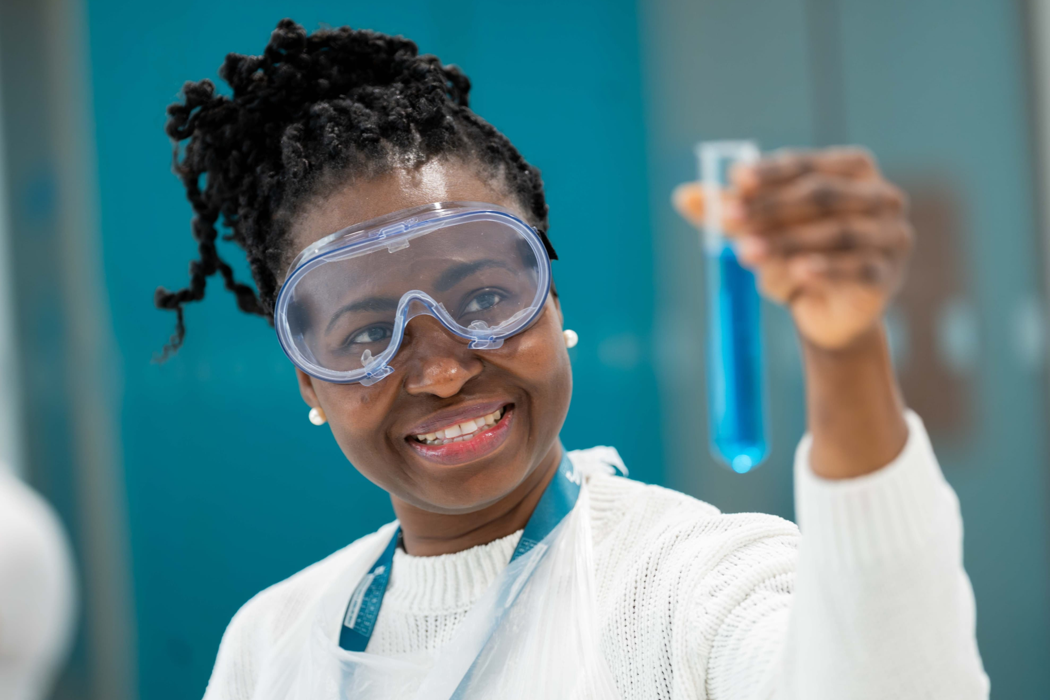 A student wearing goggles lifts up a test tube, smiling