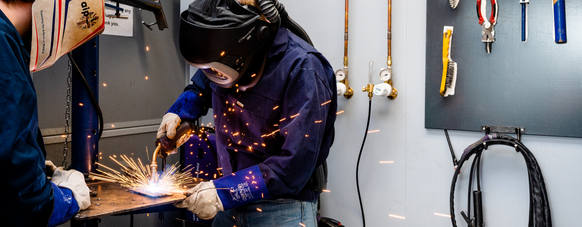 Two people wearing welding masks in a workshop, one is holding a welder while sparks fly.