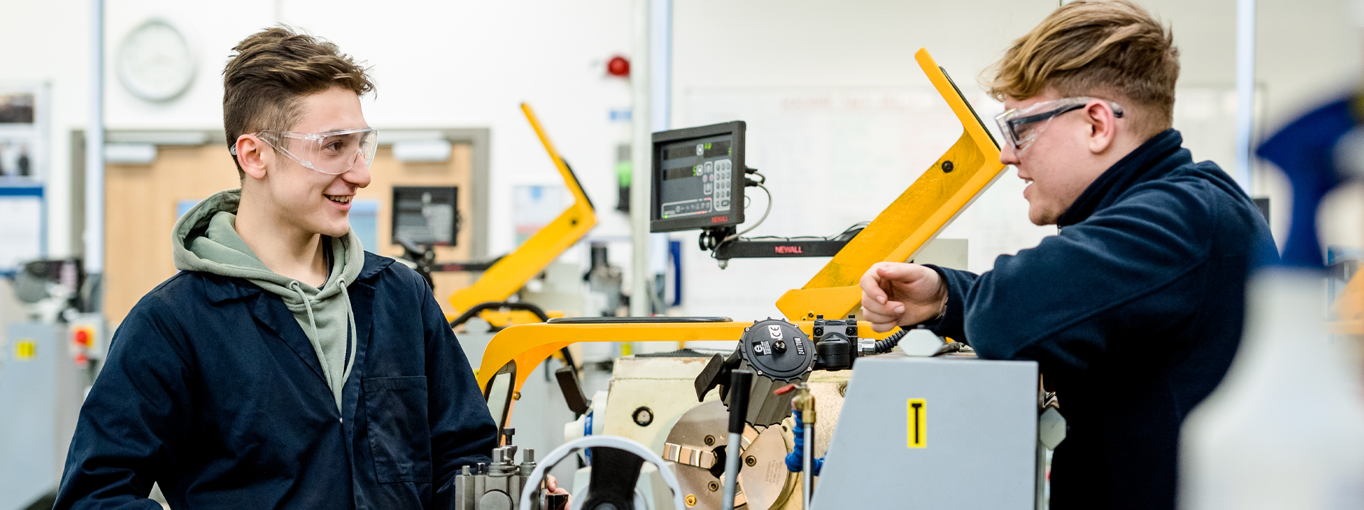 Two male students wearing safety goggles work together on a machine in a workshop.