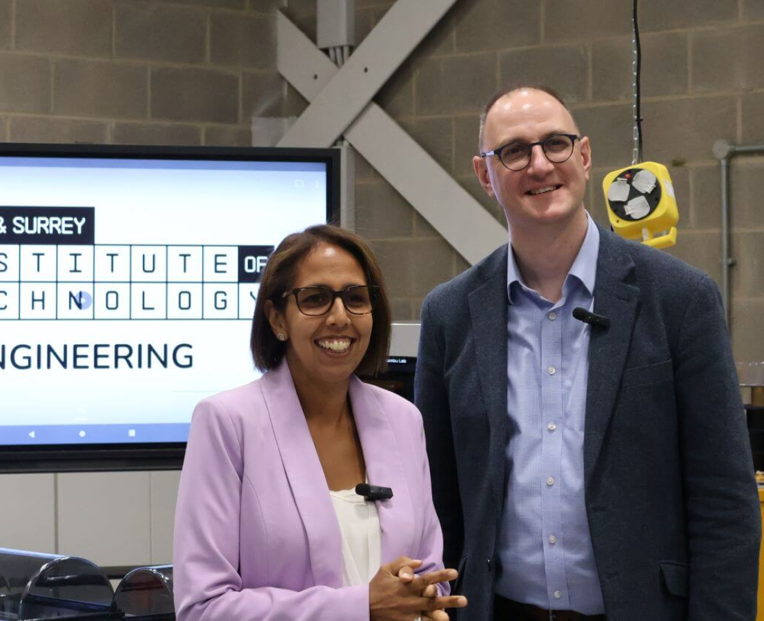 Two people stand in front of a screen welcoming them to Sussex and Surrey IoT, smiling