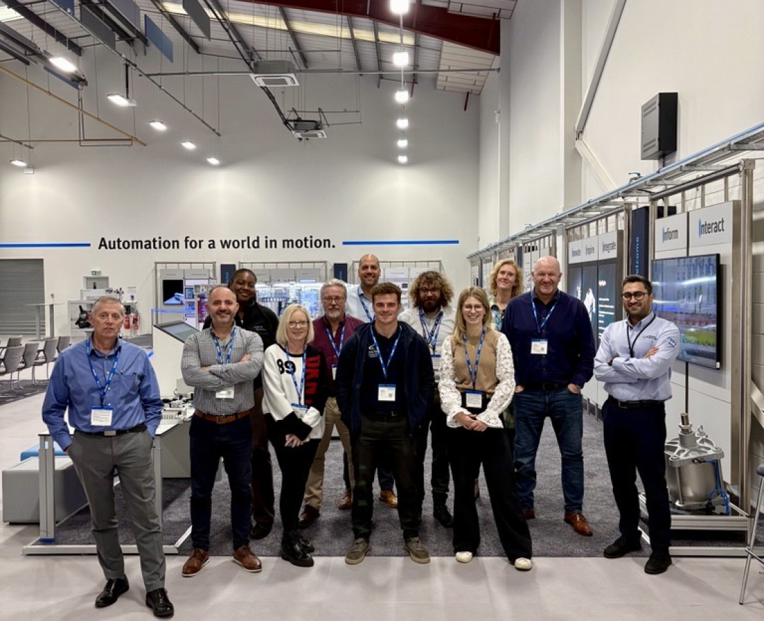 A group of smiling people in professional clothing and wearing lanyards stand in a manufacturing workshop
